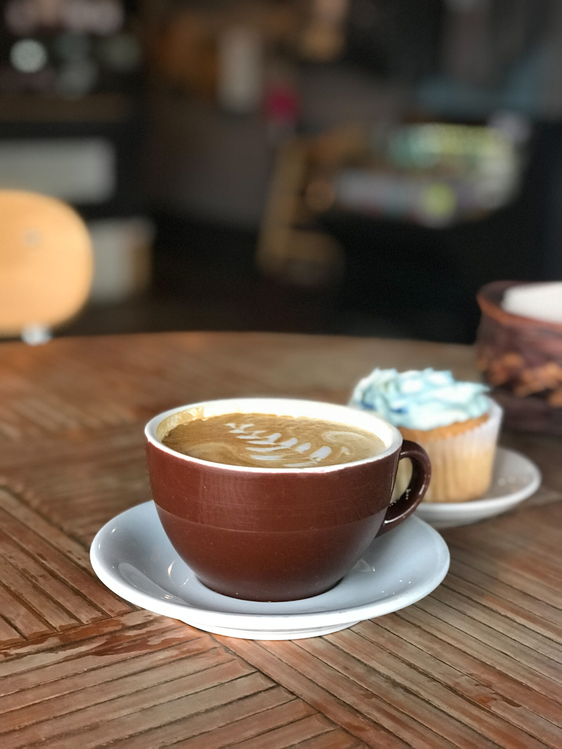 large coffee latte and cupcake on a round wooden table with coffee shop in background